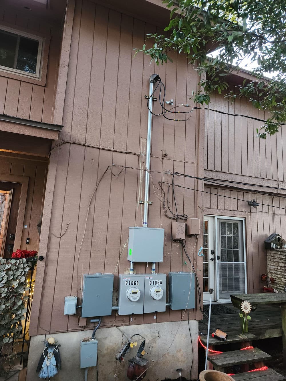 Electrical meters and service panel on a brown residential exterior wall with wiring visible.