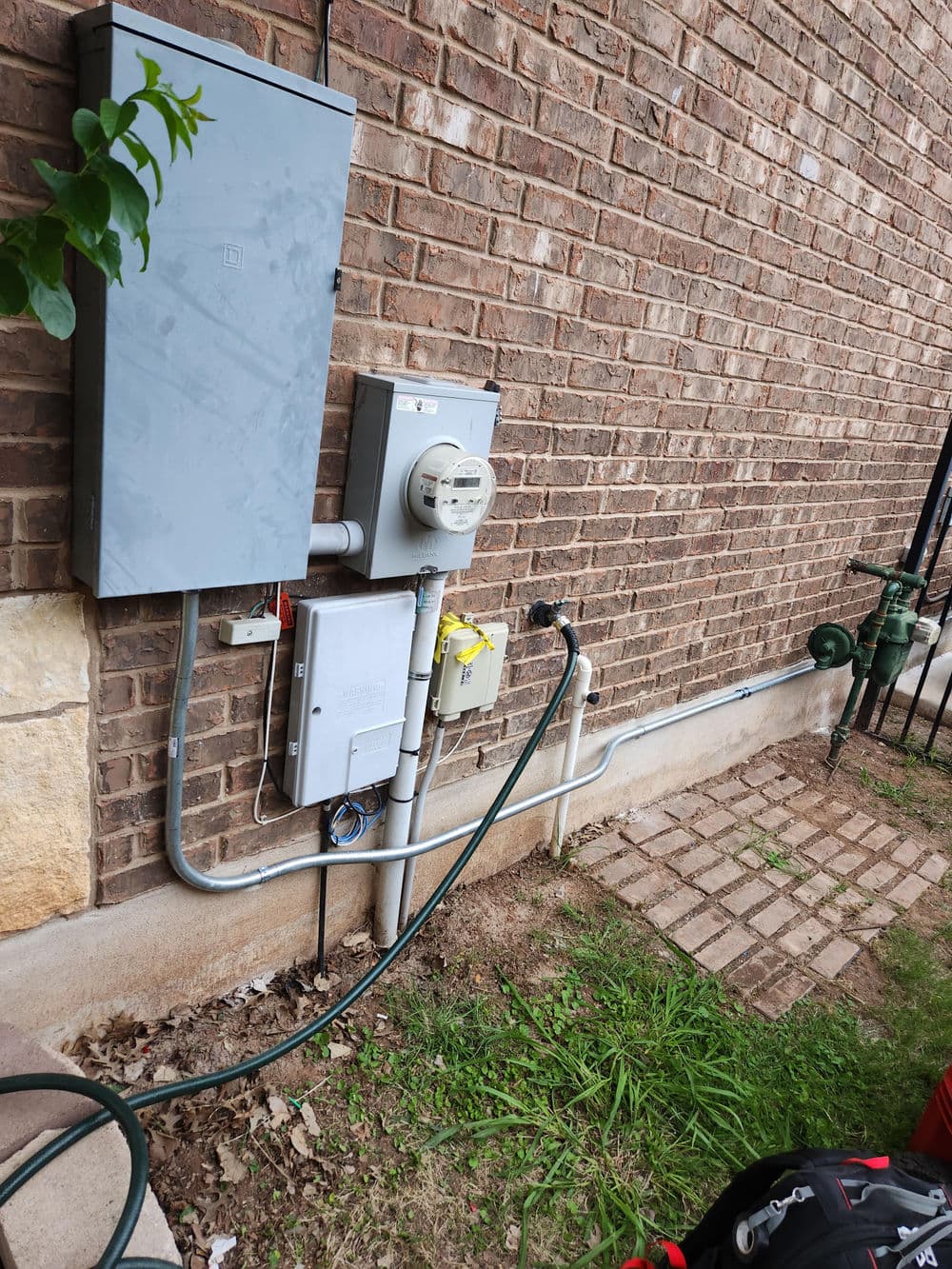 Electrical meter and service panel mounted on a brick wall beside a hose connection.