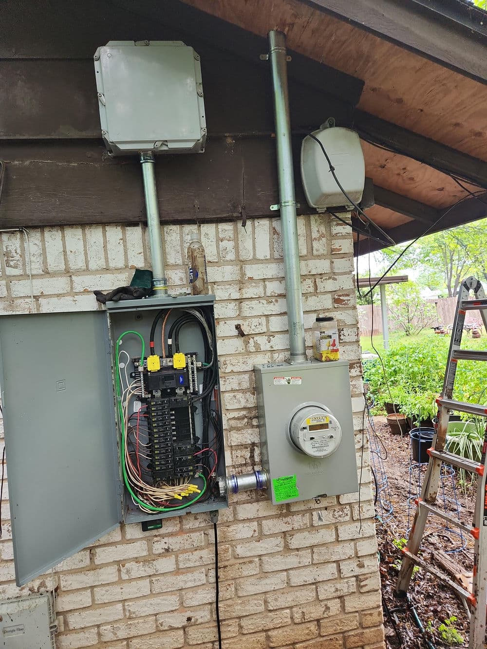 Electrical panel and meter installation on a brick wall, with wiring and nearby ladder.