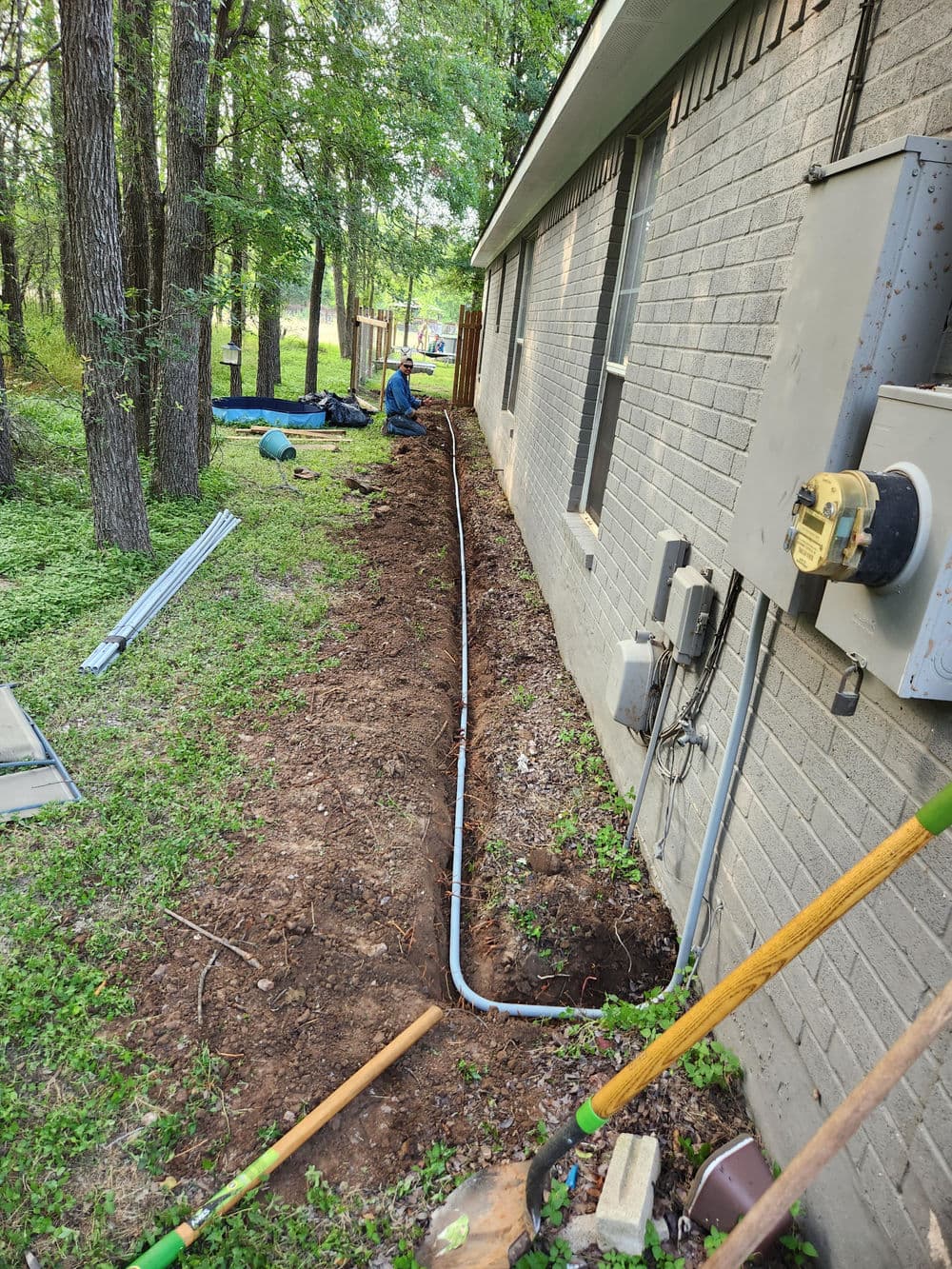 Worker installing a drainage pipe along the side of a house in a grassy yard.