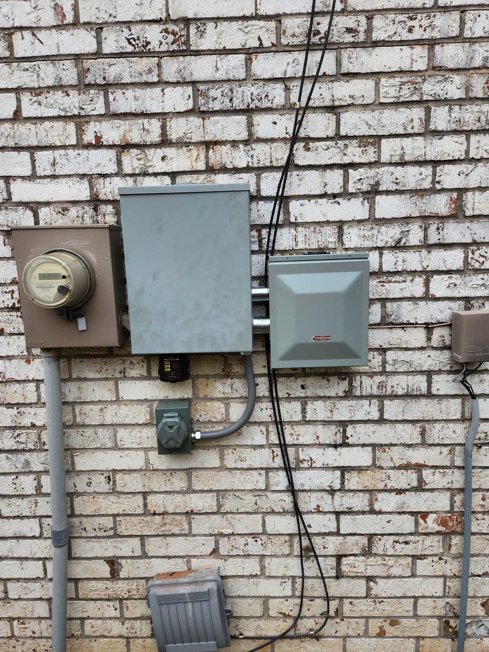 Electrical meter and service panel mounted on a brick wall with wiring connections.