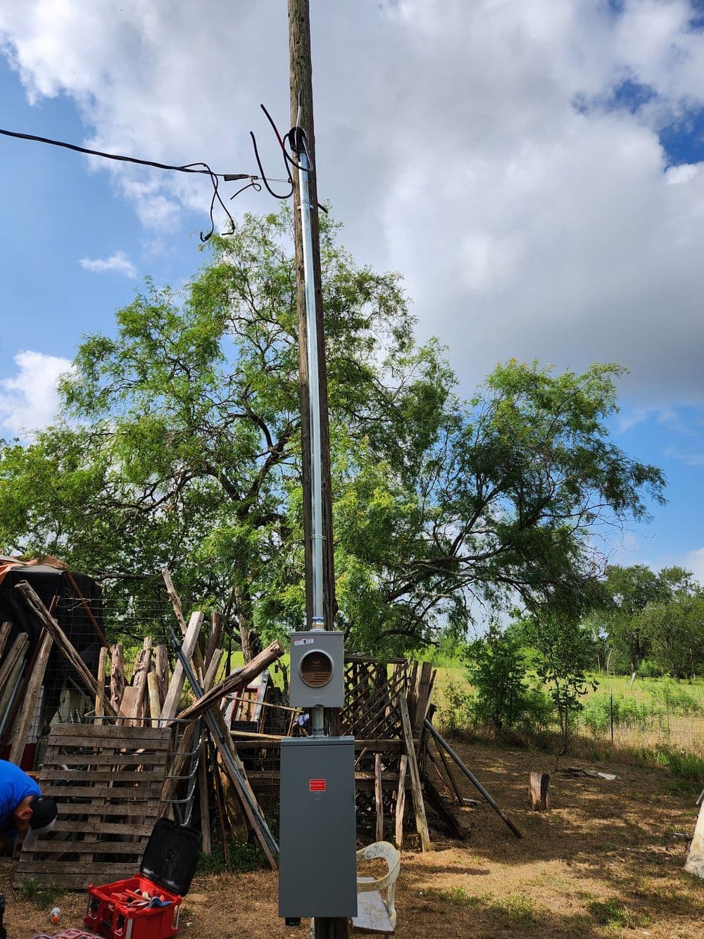 Utility pole with electrical meter box under a cloudy sky and surrounding trees.