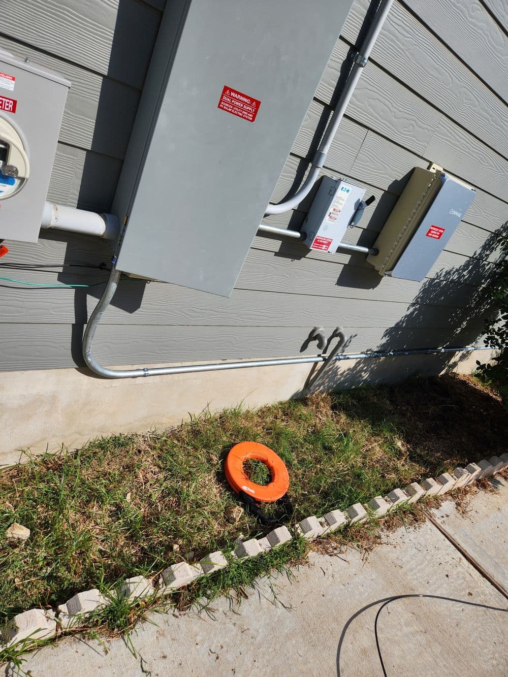 Utility meters and orange safety ring by exterior wall, with grass and concrete pathway.