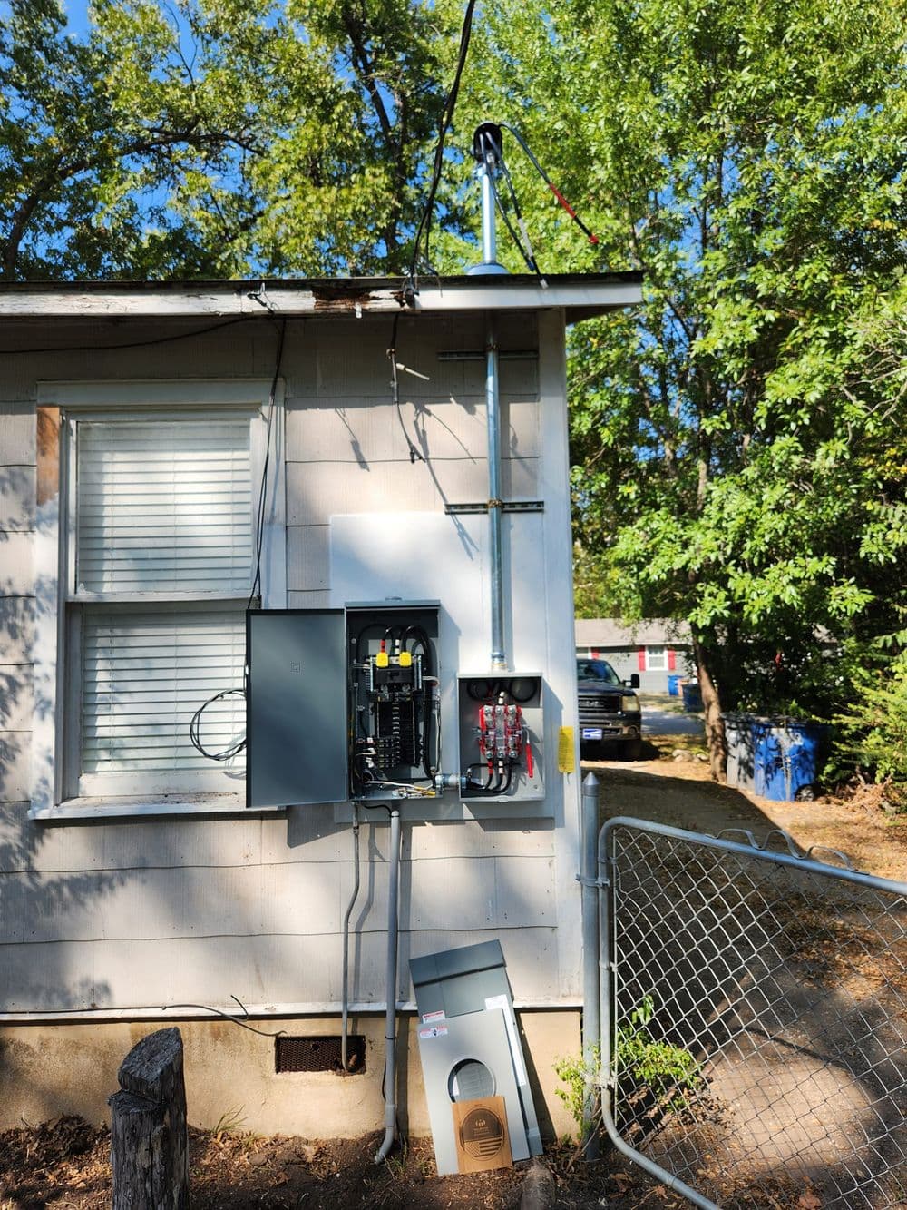 Electrical panel mounted on house exterior with nearby antenna and gated access.